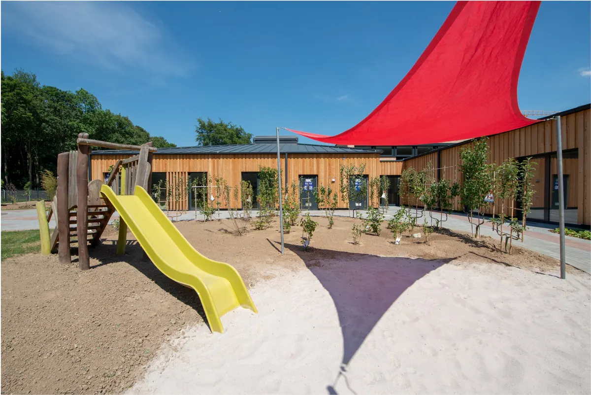 Spielbereich mit Sand, gelber Rutsche und rotem Sonnensegel vor der Holzfassade