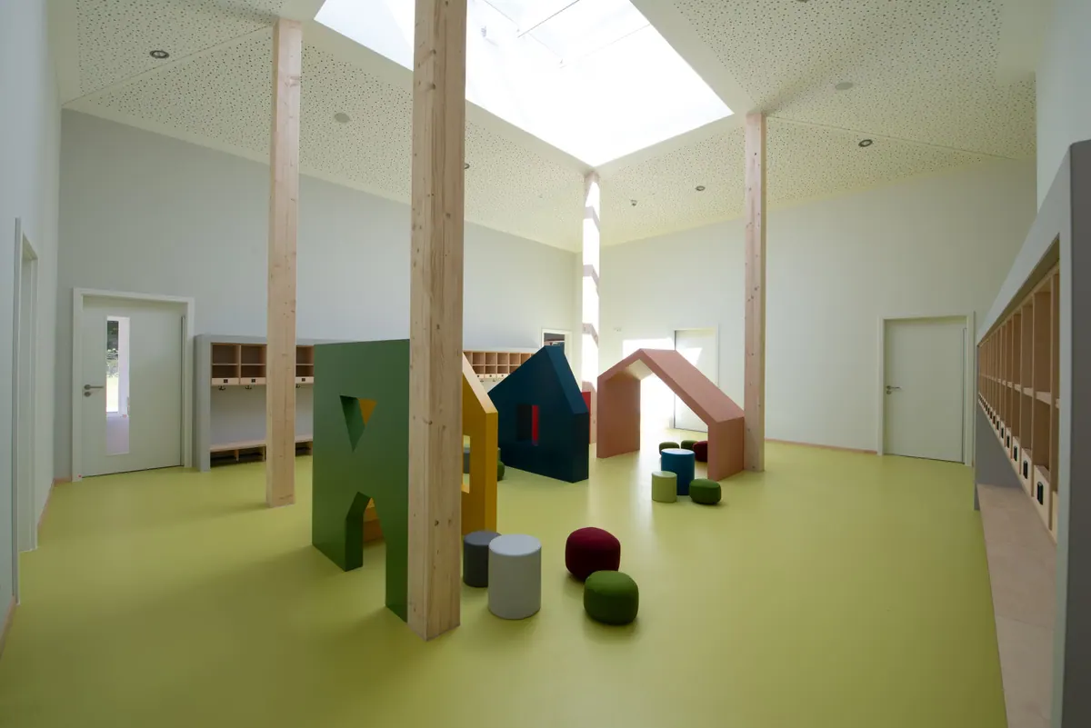 Heller Bewegungsraum mit grünem Boden, Oberlicht, Holzstützen und kleinen Spielhäusern