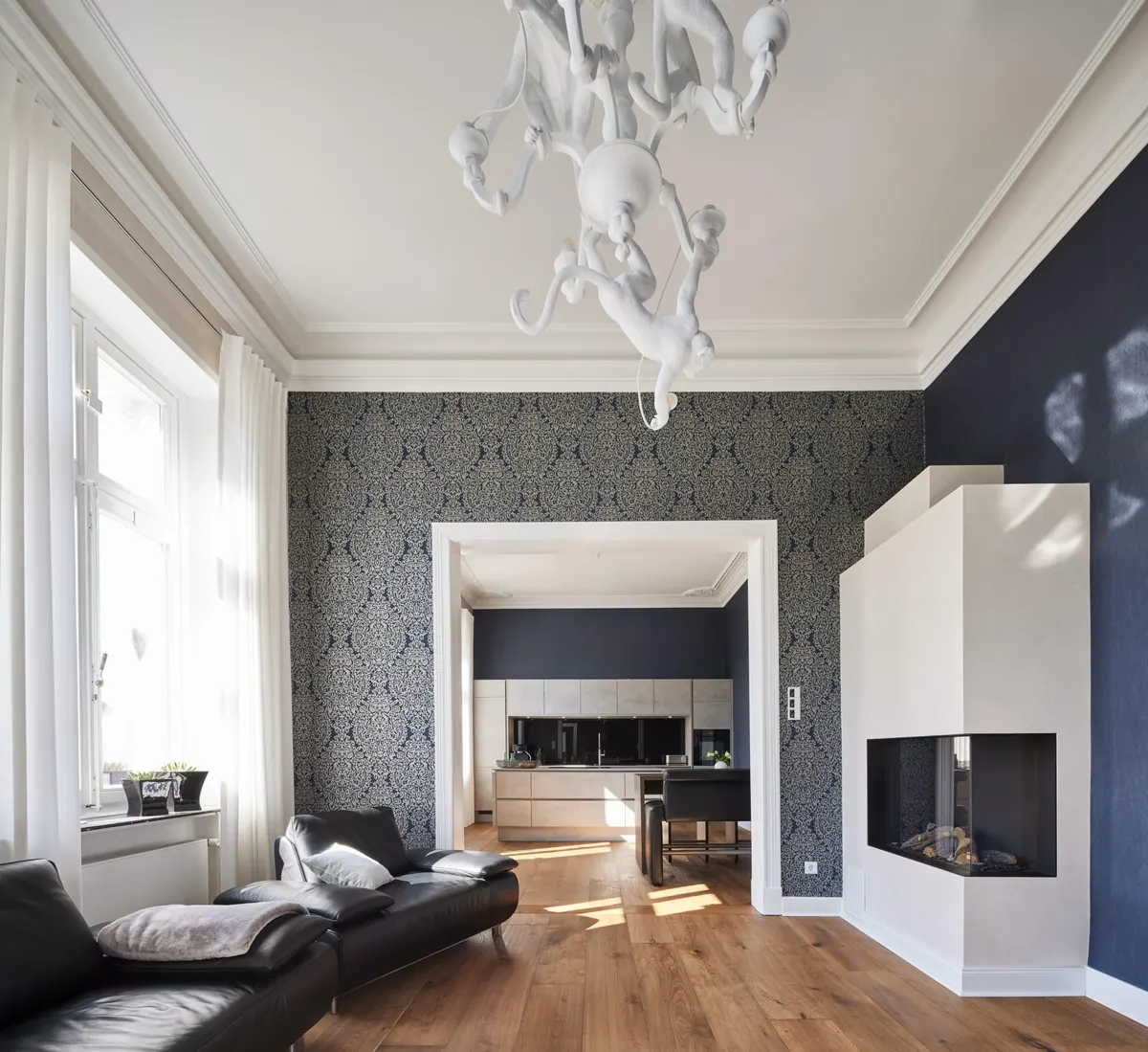 Woonkamer met stucornamenten aan het plafond, schouw, donkere bank en doorkijk naar de keuken