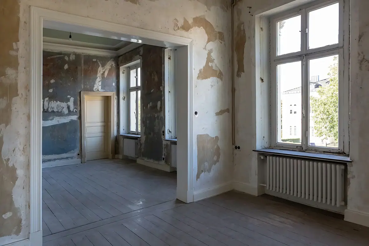 Voor de renovatie: kamer met afgebrokkeld pleisterwerk, oude vloerdelen en hoge ramen